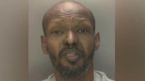 A mugshot of Mohammed Ibrhim looking at the camera. He is bald, has a black beard and goatee, with grey hairs.
