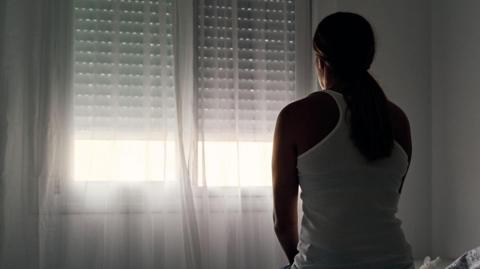 A woman sat staring out of a window. The blinds and curtains are mostly drawn with only a small bit of light coming through the bottom. 