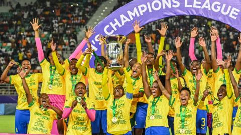 Mamelodi Sundowns players, all wearing matching yellow t-shirts and with gold medals around their necks, stand with their arms raised on a podium as  captain Zanele Nhlapho lifts the Caf Women's Champions League trophy