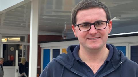 Matt Day stands beneath new white, all-weather canopy at pool wearing glasses and a blue top with a zip 