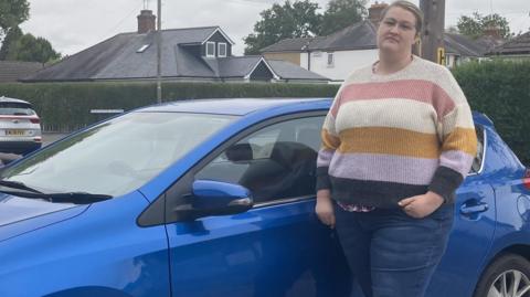 Mary Bates is standing in front of her blue car outside her home. 