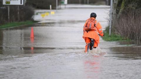 A man wearing orange wet weather gear riding his cycle through surface water on a road.