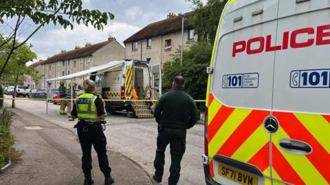 Police officers and firefighters standing in a road, next to houses, that has been sealed off with tape. The firefighters are standing next to a fire service incident vehicle and there is a police van in the foreground