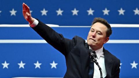 Elon Musk stands at a podium. His right arm is stretched out from his body and angled upwards at a 45 degree angle. His palm is open and flat, facing downwards.