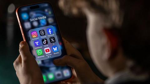 Close-up of a person holding a smartphone displaying a folder of social media apps, including Instagram, Facebook, TikTok and WhatsApp.