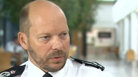 Chief Constable Rod Hansen sitting and talking to someone. He is wearing a white shirt, black tie and has black epaulettes on his shoulders.