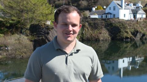 Innes MacCuidhein na sheasamh an Eilean Luinn agus gàire air aodann. 