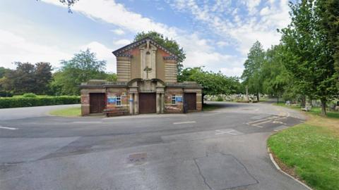 The chapel in Hull's Northern Cemetery. It is a redbrick and light stone building with a pitched roof and three large wooden doors at the front. It is surrounded by a road with trees and grass alongside and gravestones in the background.