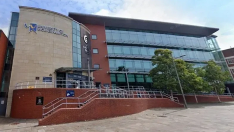 The outside of the university building. It has a glass frontage with trees in front of it on the right and steps leading up to the entrance on the left.