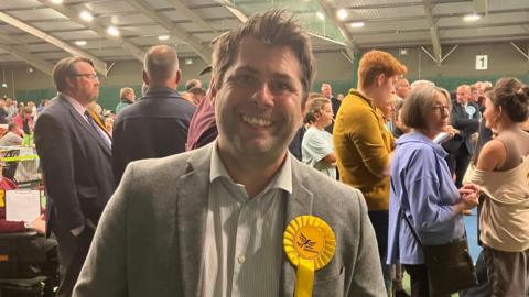 Leigh Frost wearing a grey blazer with a white striped shirt. He also has a yellow rosette on his left side with the Liberal Democrat bird logo on it. He has short brown hair and is stood in front of a crowd of people at the count.