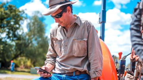 A farmer wearing a hat, sunglasses, jeans and shirt leans on a tractor and looks at a smartphone.