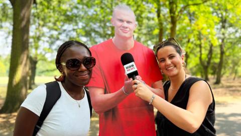 Two young women pose in front of a life-size cutout of Erling Haaland in a park.