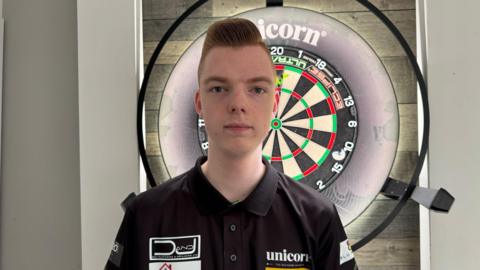Jackson looking at the camera in front of darts board. 