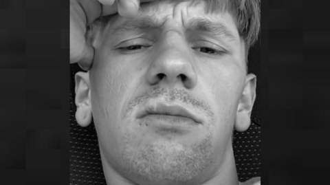 Lee Joshua Grant wears a frown in a black-and-white photo, which shows a stubbly moustache and goatee beard. His right hand touches his forehead.