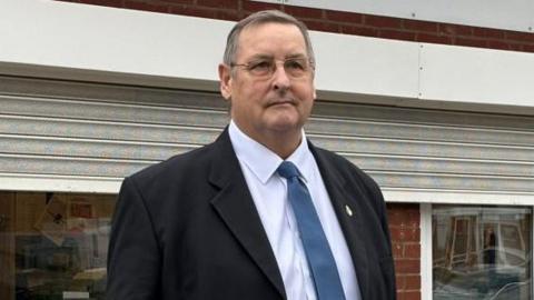 A man with short grey hair, glasses, a black jacket, blue tie and white shirt stands in front of a building with the shutters partially down.