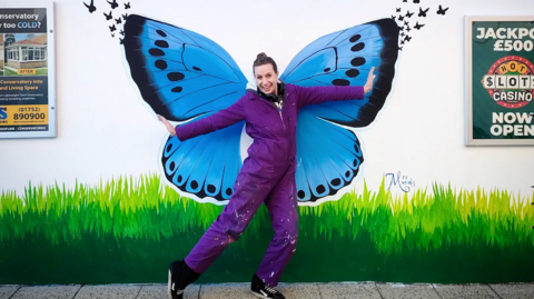 A woman dressed in a purple jumpsuit with her hair in a bun, stood in front of a painted mural she created of a blue butterfly above green grass.