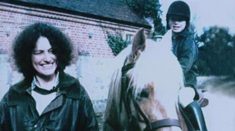 A woman with shoulder length dark curly hair and wearing a dark coloured jacket and light coloured blouse is smiling as she stands next to a young girl riding a horse. The girl is also smiling. A brick building and trees can be seen behind them.