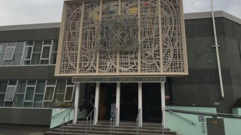 A two-storey building with a sign above the entrance reading: Plymouth Crown and County Courts. There are four entrance doors with steps leading up to them. Above the sign is a large decorative design of wrought stone work.