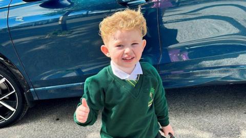 Peter Maughan, a four-year-old boy with short, blond hair, is standing by a blue car wearing a school uniform. He has a white shirt, a green and yellow tie and a green jumper. He is holding a green schoolbag and is making a thumbs-up sign.