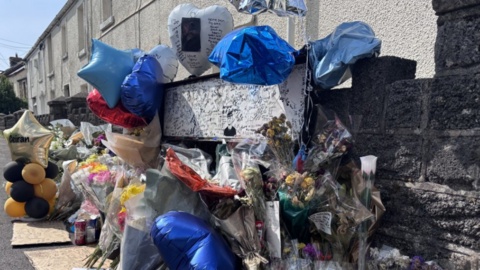 Tributes paid to Ceiran Evans, including blue balloons and banners attached to a stone wall outside a row of terrace houses. Rows of flowers are leaning against the wall.