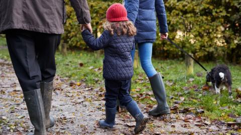 A child wearing a dark blue quilted jacket, jeans and wellies walks hand in hand with two adults on an autumnal path with leaves on the ground - they are walking a black-and-white dog 