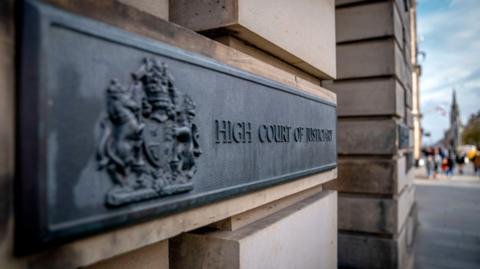 A black sign for the high court on a light sandstone with a coat of arms on the near side.