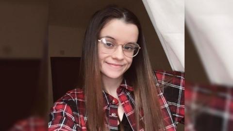 A young woman with long hair and glasses, wearing a red checked shirt.