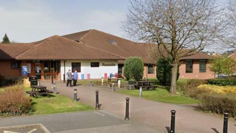 A single storey building with a white rendered wall sits back from a pathway. The building has the word Reaside on it, and two people in medical clothing are walking out of it. There are trees, hedges, and benches around the pathway