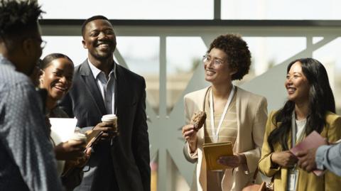 Group of young people in professional clothes standing in a group smiling and laughing, holding coffees