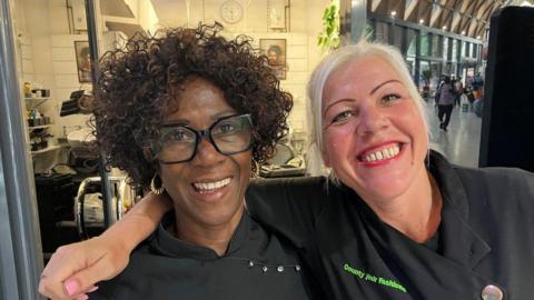 Two women are shown smiling at the camera. They are pictured from chest level. One of the women who is white with blond hair pulled back has her arm around the shoulder of the other who is black, with short curly hair and wearing black-framed glasses. They are both wearing black tunic tops.