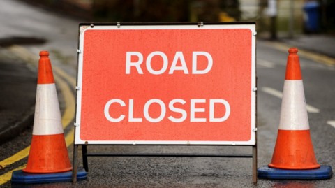 A red 'ROAD CLOSED' sign with two red and white cones on either side