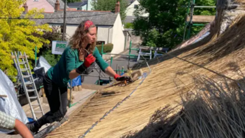 A woman with long brown hair in a green top on a roof, thatching