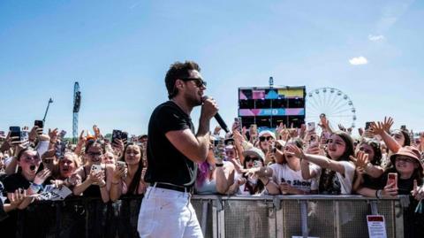 Niall Horan stands side on to a crowd. He is wearing a black t-shirt and white jeans and is wearing sunglasses. He is holding a microphone in his hand. People in the crows are holding out their arms to him and holding phones, In the background is the control tower for the stage which is partially obscuring a big wheel