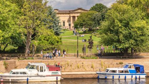 A general view of the Yorkshire Museum behind a river with two boats. There are also green trees in the foreground and pedestrians on a path.