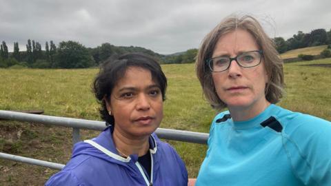 Two women stand in front of a metal gate backing on to fields