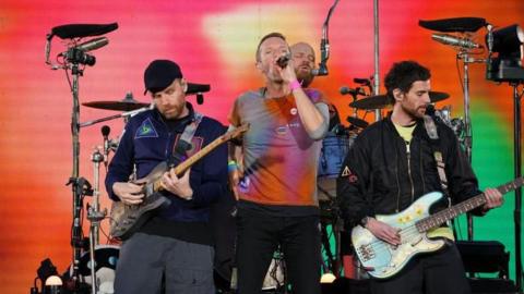 Chris Martin of Coldplay is singing on a stage, with bassist Guy Berryman playing on his right, guitarist Jonny Buckland on his left, and drummer Will Champion playing in the background. Martin is wearing an orange, blue and purple tie-dye T-shirt, with the others wearing dark-coloured jackets.