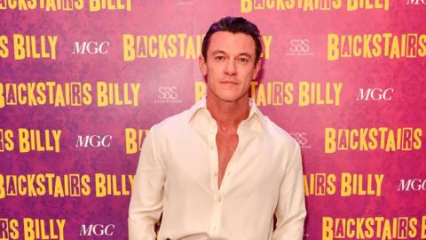 Welsh actor Luke Evans standing in front of a background that says Backstairs Billy, he is wearing a whit shirt that is unbuttoned. 