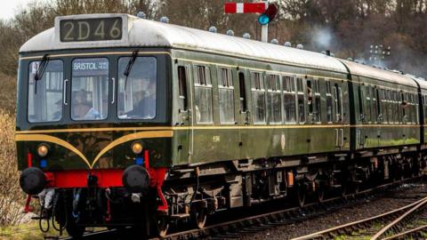 A steam locomotive that is dark green and has lots of windows. Steam is coming from funnels on top of it. A number above its cabin reads 2D46