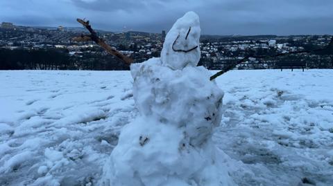 A snowman positioned in the middle of a snowy field. The snowman is roughly shaped, with an uneven body and head, and it has two stick arms extending outward, one on each side. A simple face is drawn on the snowman using dark lines, featuring two vertical marks for eyes and a curved line for a smile. The surrounding ground is covered in snow, with some patches appearing icy or slushy. In the background, there is a panoramic view of a Sheffield with numerous buildings and houses spread across a hilly landscape.
