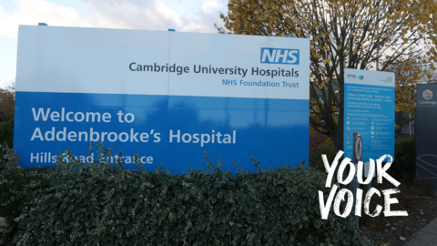 A sign at the entrance to Addenbrooke's Hospital. The top half is white with the logo of the Cambridge University Hospitals NHS Foundation Trust. The bottom half has the words "Welcome to Addenbrooke's Hospital" in white lettering on a blue background. There is a smaller blue and white sign to the right and a row of shrubs in front of the signs.
