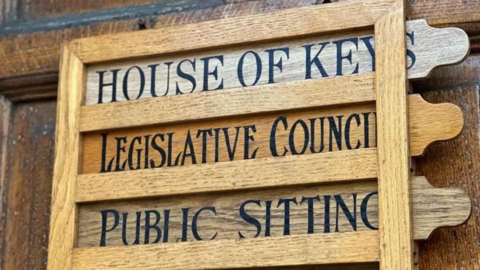 A wooden frame features three wooden slots where three wooden signs are inserted. The signs are emblazon with black, capitalised font. They say House of Keys, Legislative Council and Public Sitting.