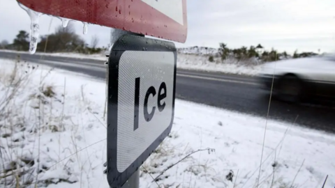 A road sign warning of ice stands on a snow-covered roadside.