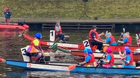 Two dragon boats, with green dragon heads and crewed by rowers in red life jackets, colourful wigs and rainbow skirts, prepare to set off on the River Ouse.