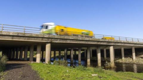 M6 Lune Gorge Castle Howe Bridge. It is a brutalist concrete structure with numerous pillars, running over a body of water with a gravelly path on one side.