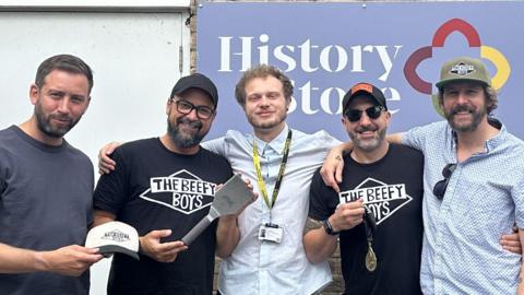 Five men posing outside a lock-up marked History Store. Three of the men hold up a cap, spatula and medal. Two are wearing black t-shirts with The Beefy boys slogan on it. A man in the centre wearing a council lanyard in a pale blue shirt has his arms around the men either side of him.