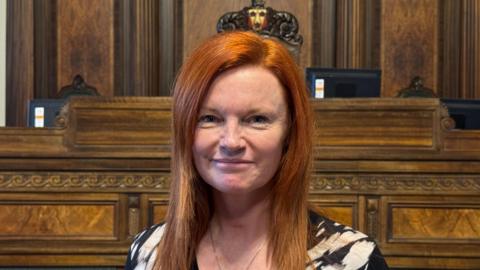 An image of a red-headed Ella Worthington, she has long hair and is smiling. She is standing in the council chamber, with wood panelling and seating behind her. She is wearing a black and white top and a necklace.
