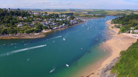 The estuary in Salcombe with boats in the water and homes overlooking the  water on a bright sunny day. Some sandy beaches are visible on the left hand side of the photograph and the river is a bright blue turquoise colour.