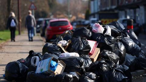 A large pile of black bin bags, with a few cardboard boxes and other bits of rubbish interspersed. The pile appears to be partly on the pavement and partly on the road. 