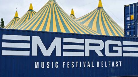 Five yellow and blue striped teepee tents behind a blue cargo storage shed with big bold white letters on it that say 'EMERGE' MUSIC FESTIVAL BELFAST in white.