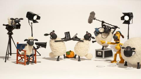 Shaun the Sheep sits in a director's chair with a megaphone while other sheep act out a scene around him